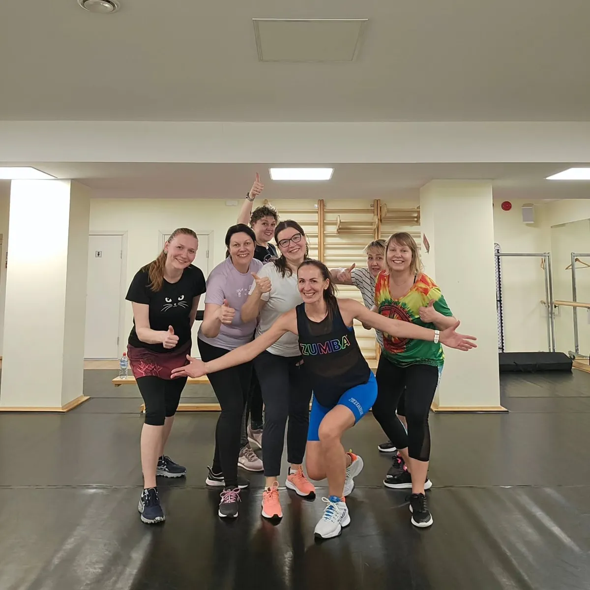 Sanita ar izplestām rokām priekšā, aiz viņas smaidošas dalībnieces Zumba nodarbības zālē